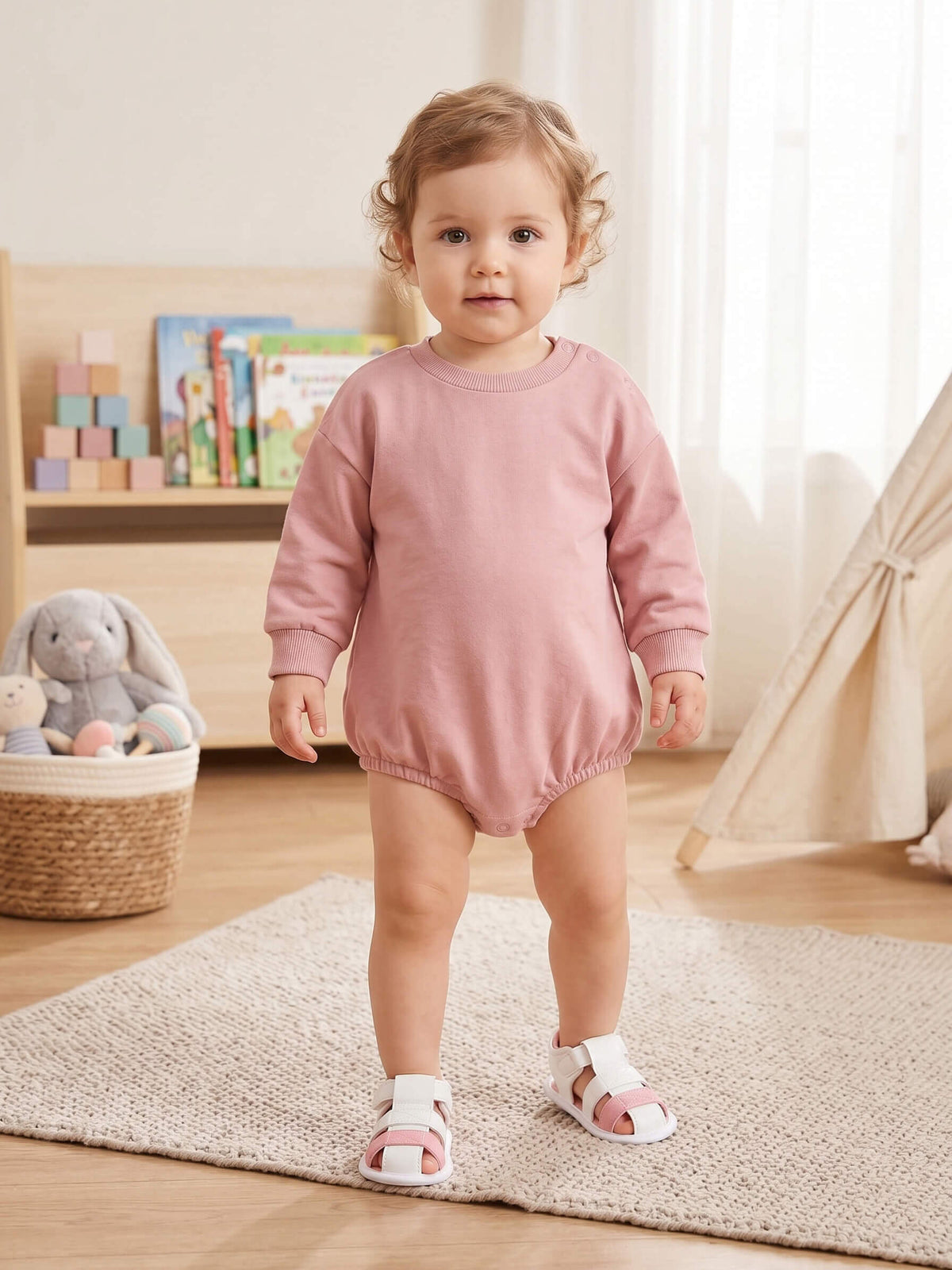 Baby in a pink outfit standing on a rug in a room with toys and books.